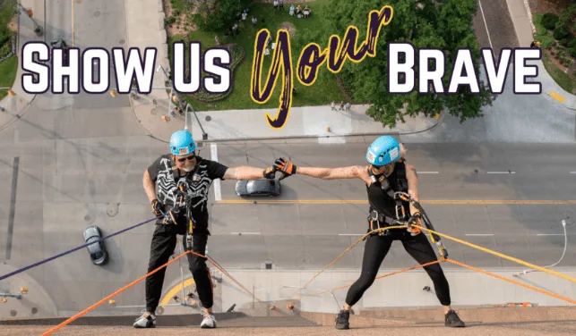 Two people in helmets and harnesses rappelling down the side of a downtown Windsor building under the words Show Us Your Brave.