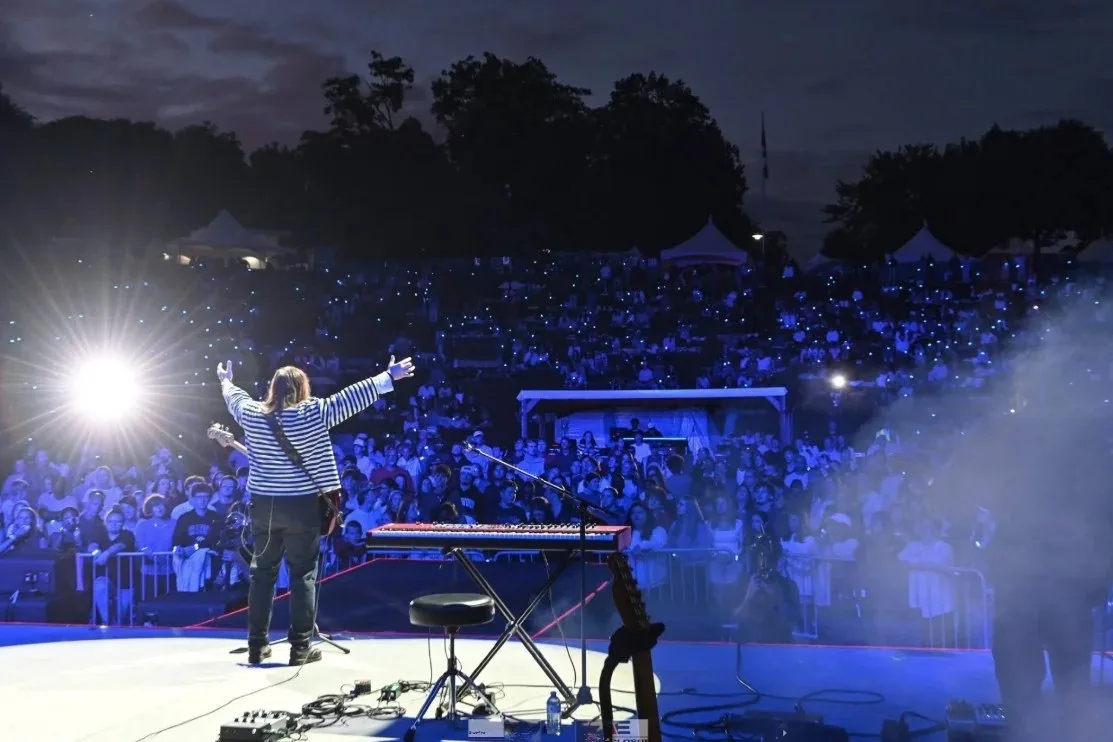 A musician on stage with arms raised, facing a large outdoor evening crowd lit blue by stage lights.