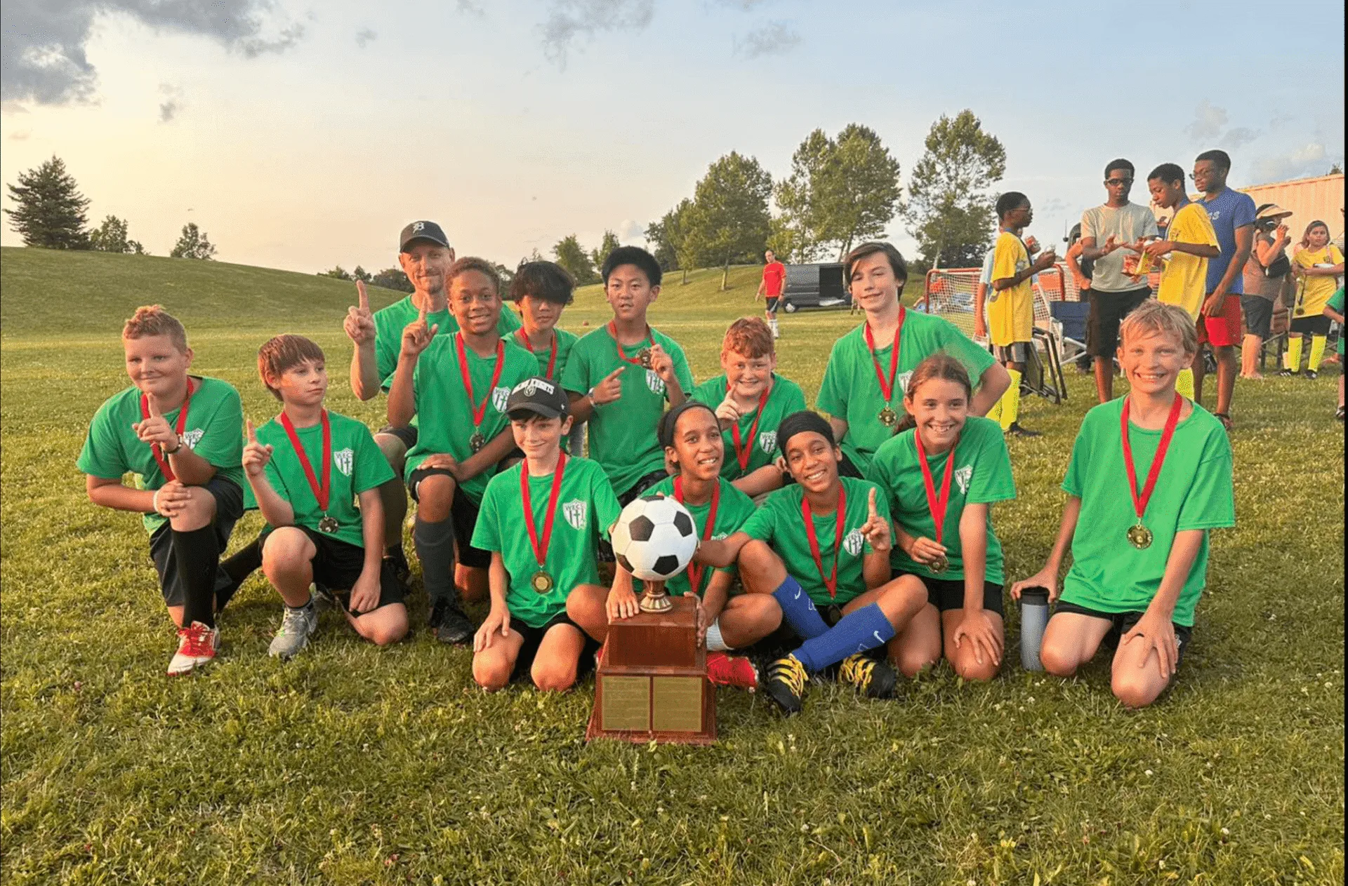 A youth soccer team in green kit kneeling on a grass pitch behind a trophy and a ball, wearing medals.