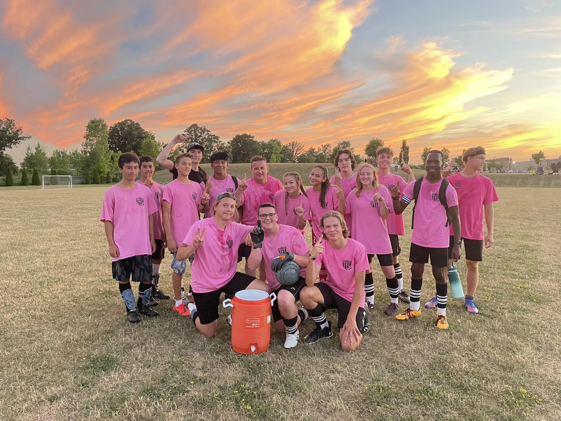 A youth soccer team in pink shirts gathered on a grass field around an orange water cooler at sunset.