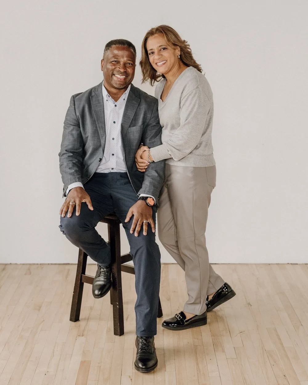 Des and Kim Griffiths in a studio portrait, Des seated on a stool and Kim standing beside him with her arm linked through his.