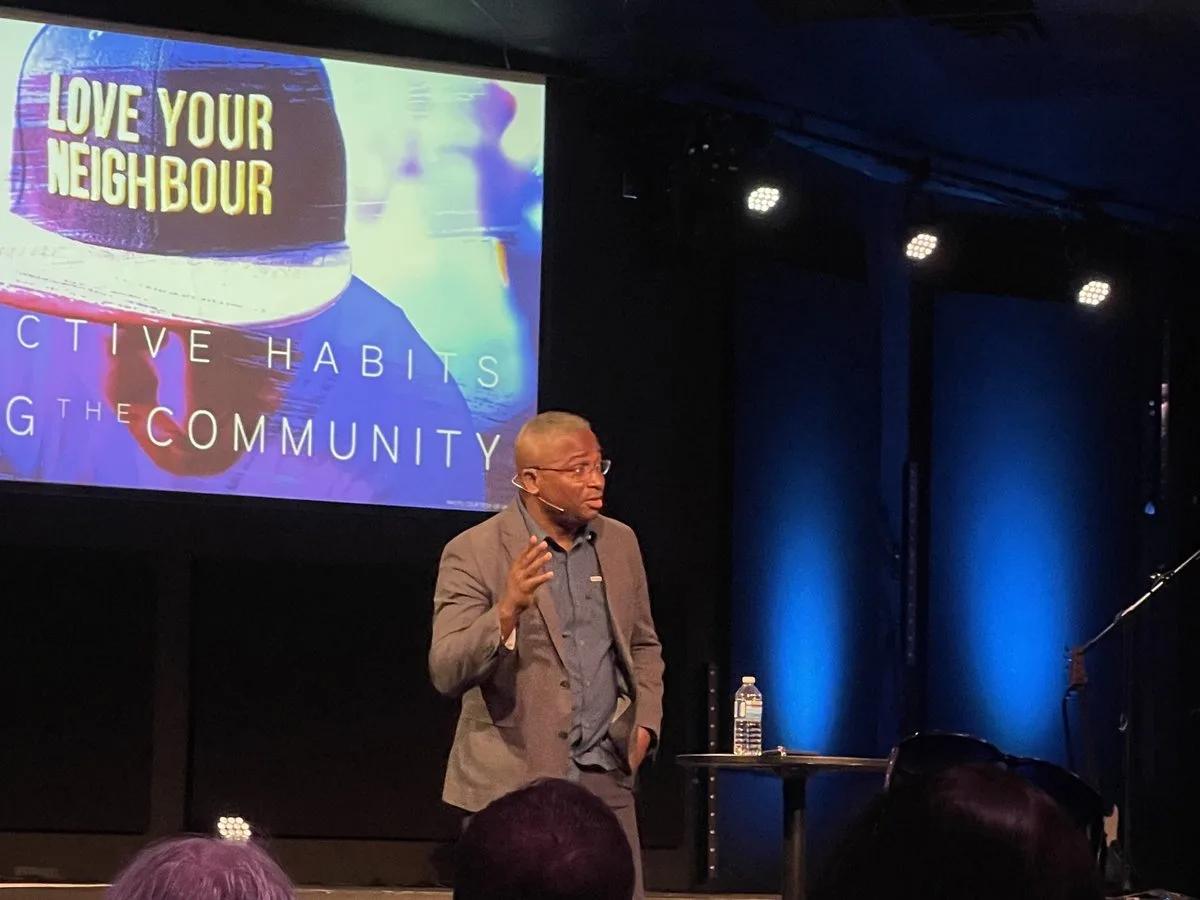 Des Griffiths speaking on stage in front of a slide reading Love Your Neighbour, with audience members in the foreground.