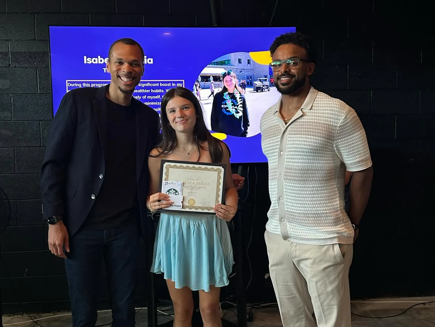 A young participant holding her program certificate between two mentors, with a presentation slide behind them.