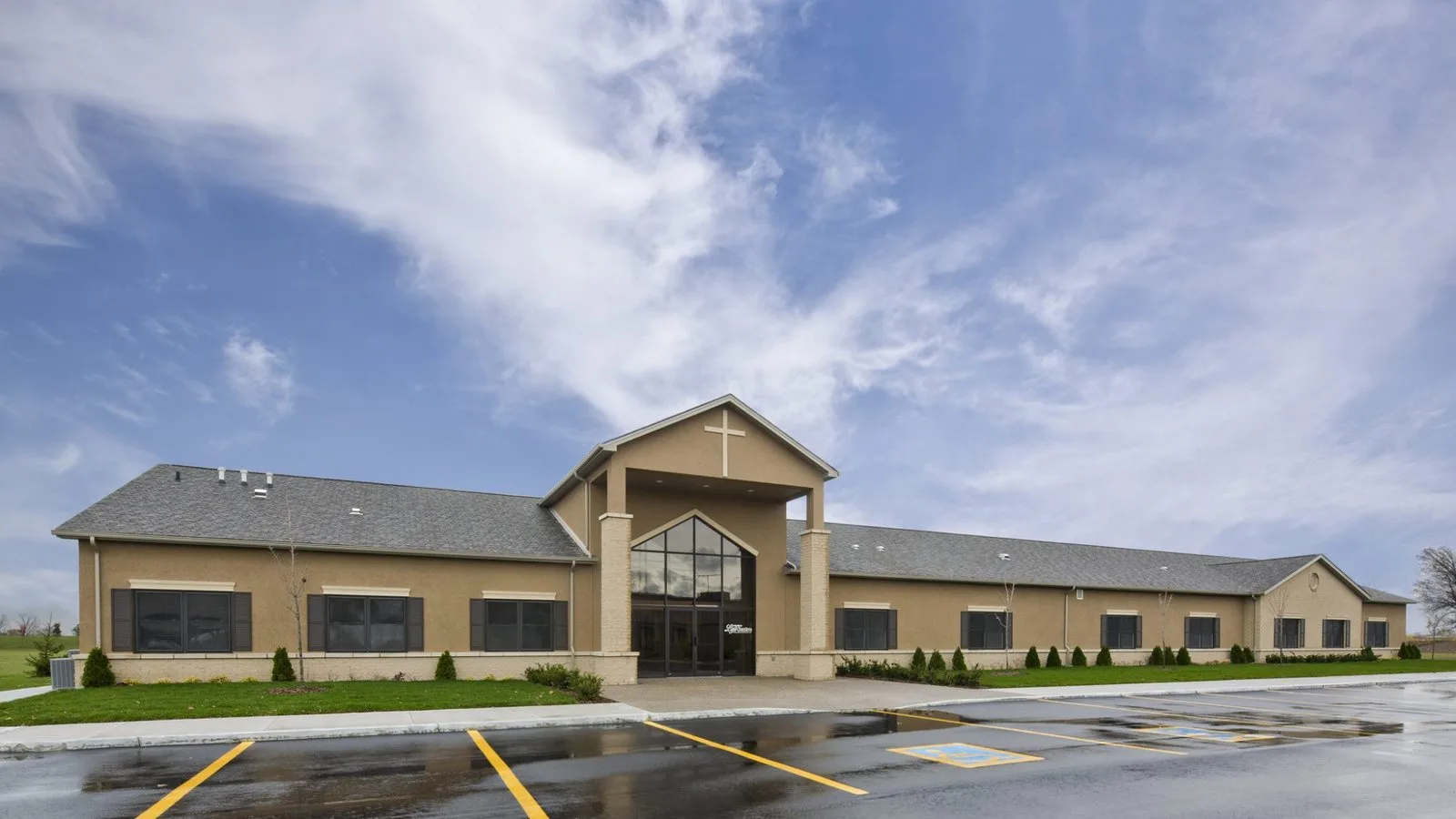 Exterior of a tan single-storey building with a peaked, glass-fronted entrance topped by a cross.